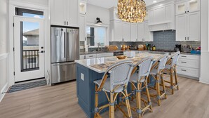 Une cuisine moderne avec des meubles blancs, un grand îlot central doté de chaises, un lustre et un réfrigérateur en acier inoxydable.