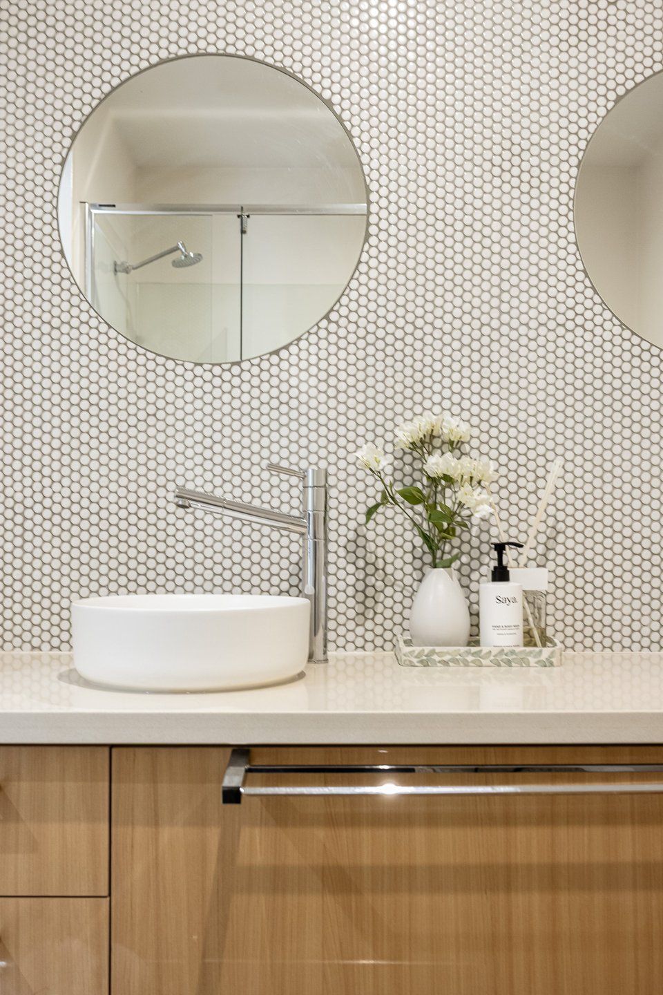 A bathroom with a white sink, two round mirrors, a modern faucet, a vase with flowers, and a bottle of soap.