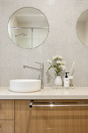Een badkamer met een witte wastafel, twee ronde spiegels, een moderne kraan, een vaas met bloemen en een zeepfles.