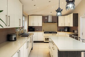 A modern kitchen with white cabinets, a central island, stainless steel appliances, and a decorative chandelier. - Spacious 4BR Home with Garden and Parking (London)