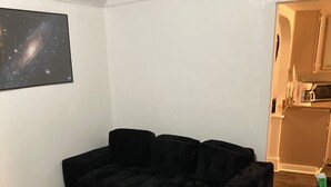 A room with a black sofa, a small table, a kitchen area with cabinets, and a framed picture on the wall.