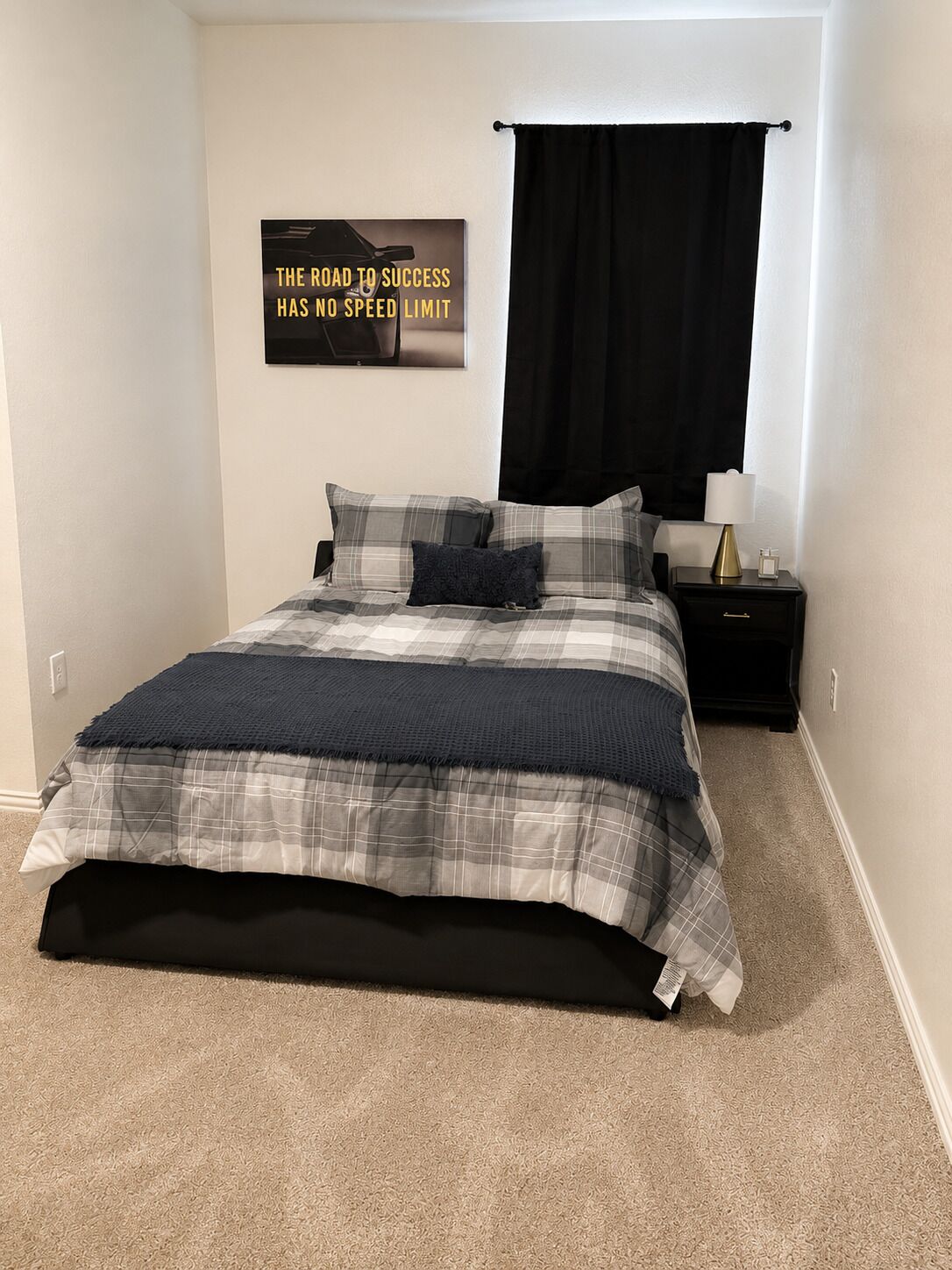 A bedroom with a bed, a nightstand, a lamp, a black curtain, and a motivational poster on the wall.
