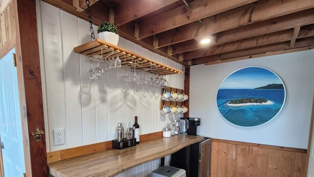 A kitchen with wooden beams, a bar area with stools, a hanging wine rack, and a wall-mounted painting of a coastal scene.