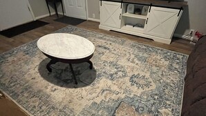 Un salon avec une table basse ronde, un tapis à motifs, un téléviseur et un meuble blanc.
