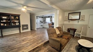 Un salon avec un canapé, un fauteuil et une petite table. Au fond, on aperçoit une cuisine équipée de placards et d’un four à micro-ondes.