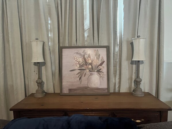 A framed artwork of flowers on a wooden table with two lamps, set against a backdrop of curtains.