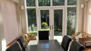 Ein Essbereich mit Tisch und Stühlen, ein Korbstuhl und Blick durch Glastüren in einen Garten.