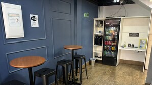 A room with two wooden tables and stools, a refrigerator stocked with beverages, and a wall-mounted sign indicating Wi-Fi availability.