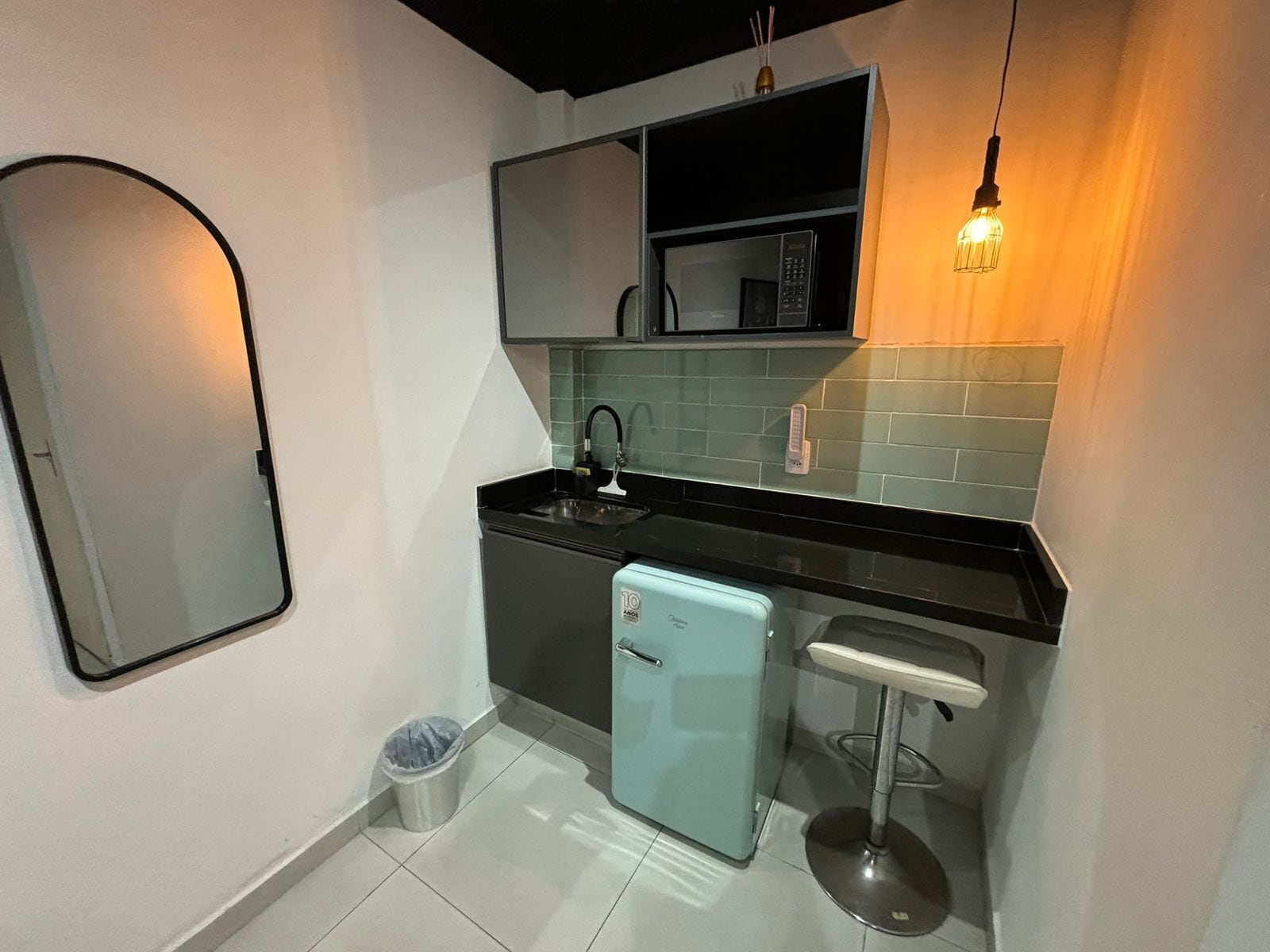 Una cocina compacta con un lavamanos, un refrigerador pequeño, un bote de basura y un taburete de bar.