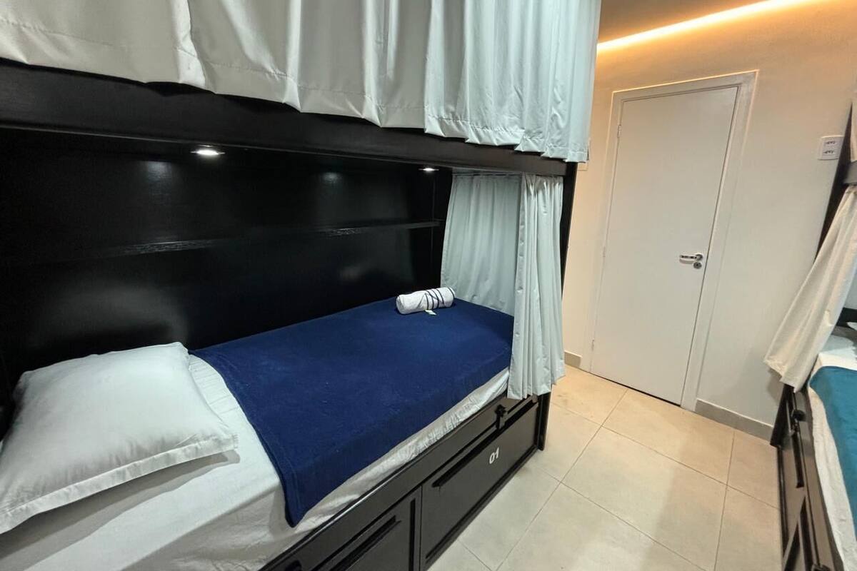 Cama litera con colchón azul, almohadas blancas y cortina blanca. Hay una puerta blanca y un pequeño armario con cajones.
