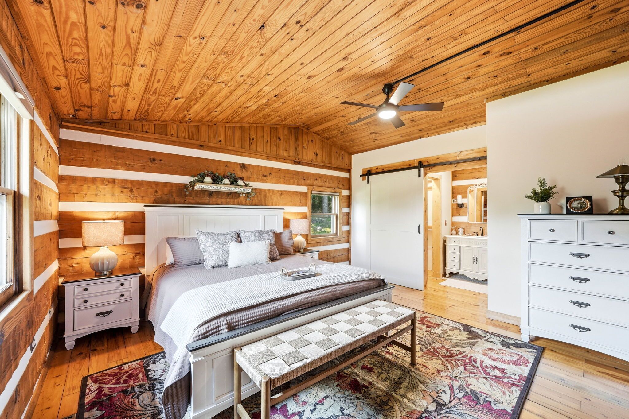 Ein Schlafzimmer mit Holzboden, einem Bett mit Sitzbank im Schachbrettmuster, einem Nachttisch, einer Kommode und einem Deckenventilator.