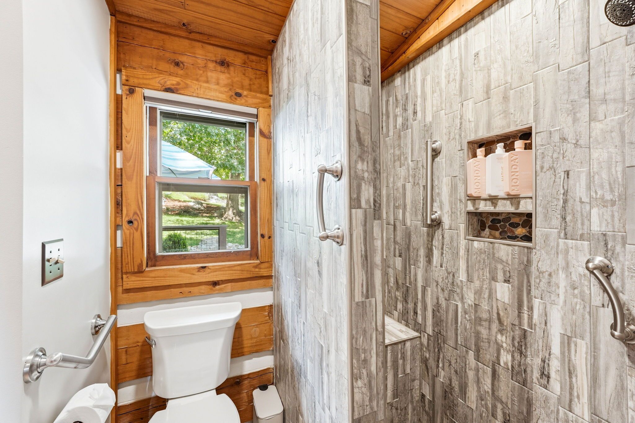 A bathroom with a wooden ceiling, a window, a toilet, and a shower with stone tiles.