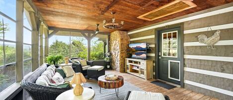 A covered outdoor seating area with wicker furniture, a stone pillar, a television, and a rooster decoration.
