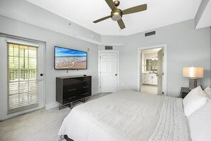 Une chambre à coucher comprenant un lit, un téléviseur, un ventilateur de plafond, une commode et une salle de bain visible à travers une porte ouverte.