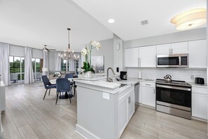 A modern kitchen with white cabinetry, a central island, and a dining area with chairs. - Resort Condo 2BR/2BA Minutes to Disney, Pool (Orlando)
