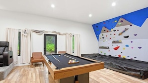 Une table de billard avec un mur d’escalade et un canapé dans une pièce donnant sur un espace verdoyant.