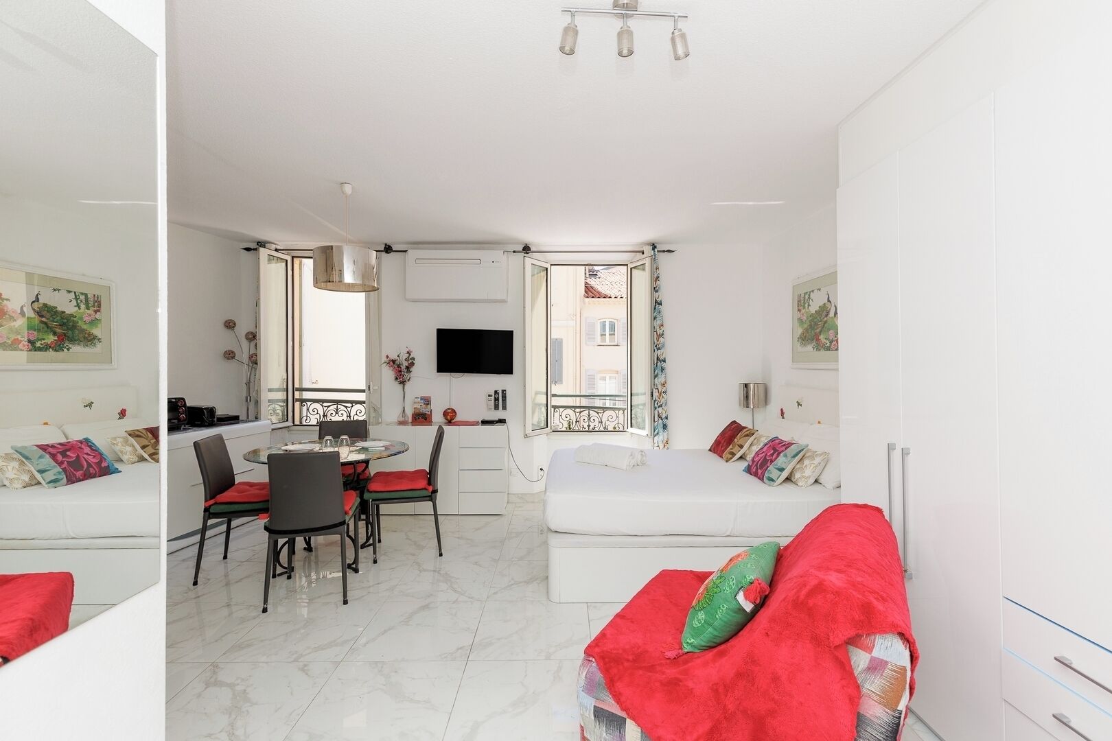 Una habitación moderna y bien iluminada con comedor, una cama con ropa de cama roja y un televisor montado en la pared.