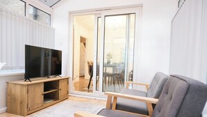 Een moderne woonkamer met een flatscreen televisie op een houten standaard, een grijze bank en een glazen deur die leidt naar een eetgedeelte.