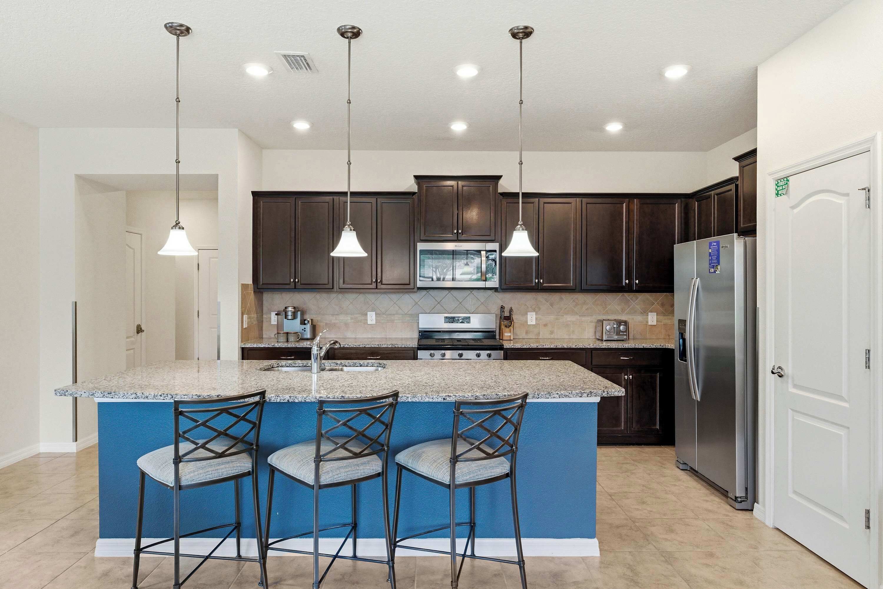 A kitchen with dark wood cabinets, a stainless steel refrigerator, a built-in microwave, and a granite island with bar stools.