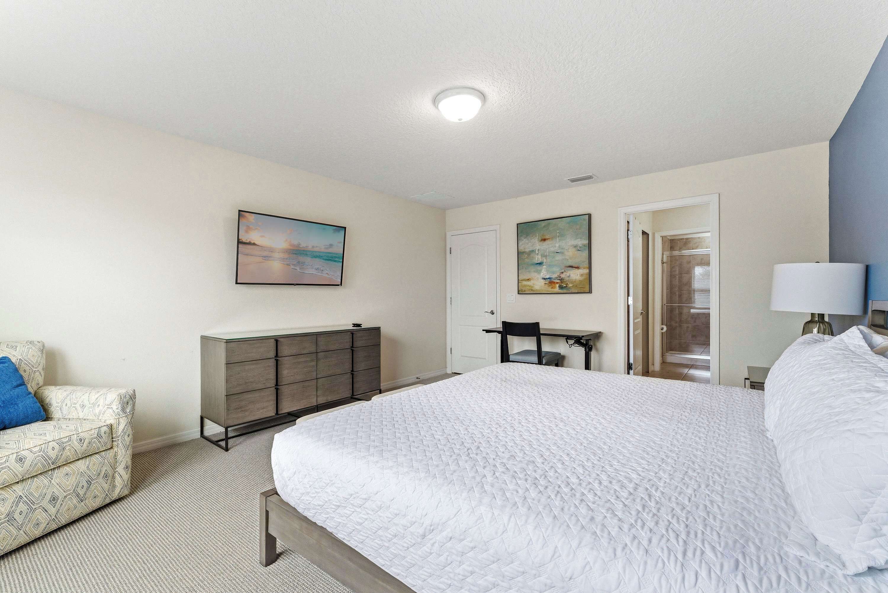 A bedroom with a bed, a dresser, a chair, a desk, and a door leading to a bathroom.