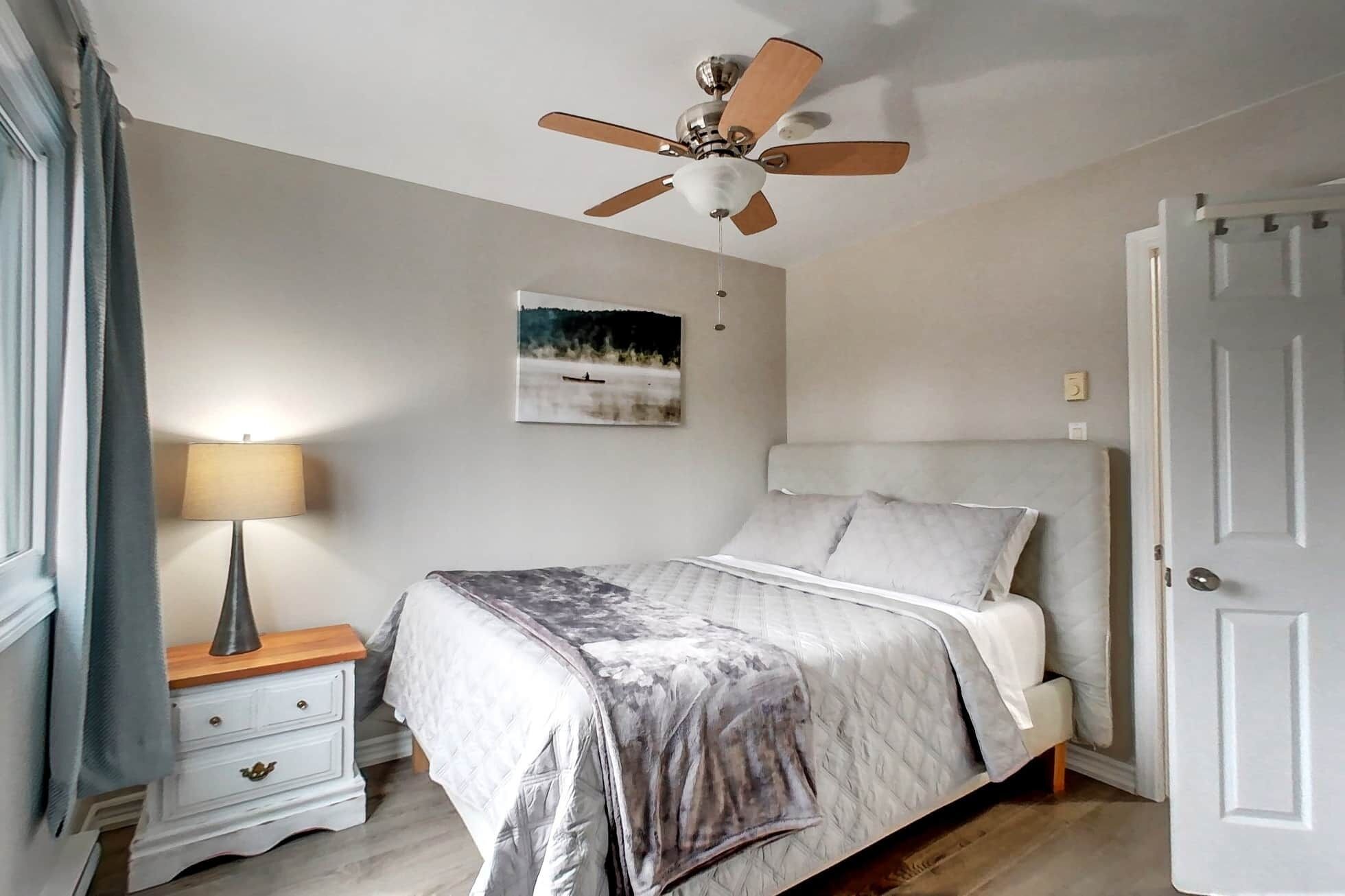 A bedroom with a bed, a nightstand, a lamp, a ceiling fan, and a door.