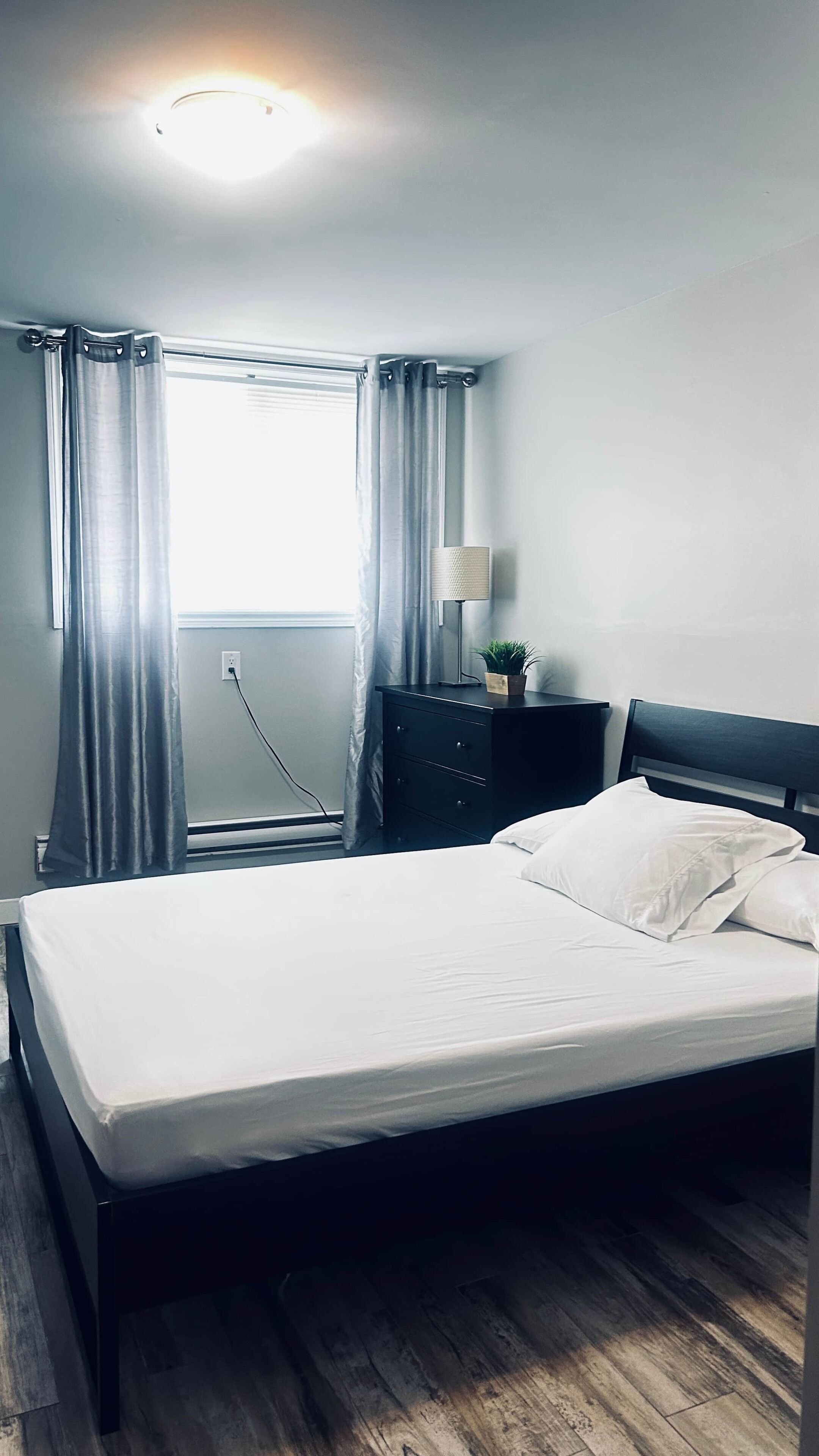 Ein Schlafzimmer mit einem Bett, einem Nachttisch, einer Lampe und einem Fenster mit Vorhängen.