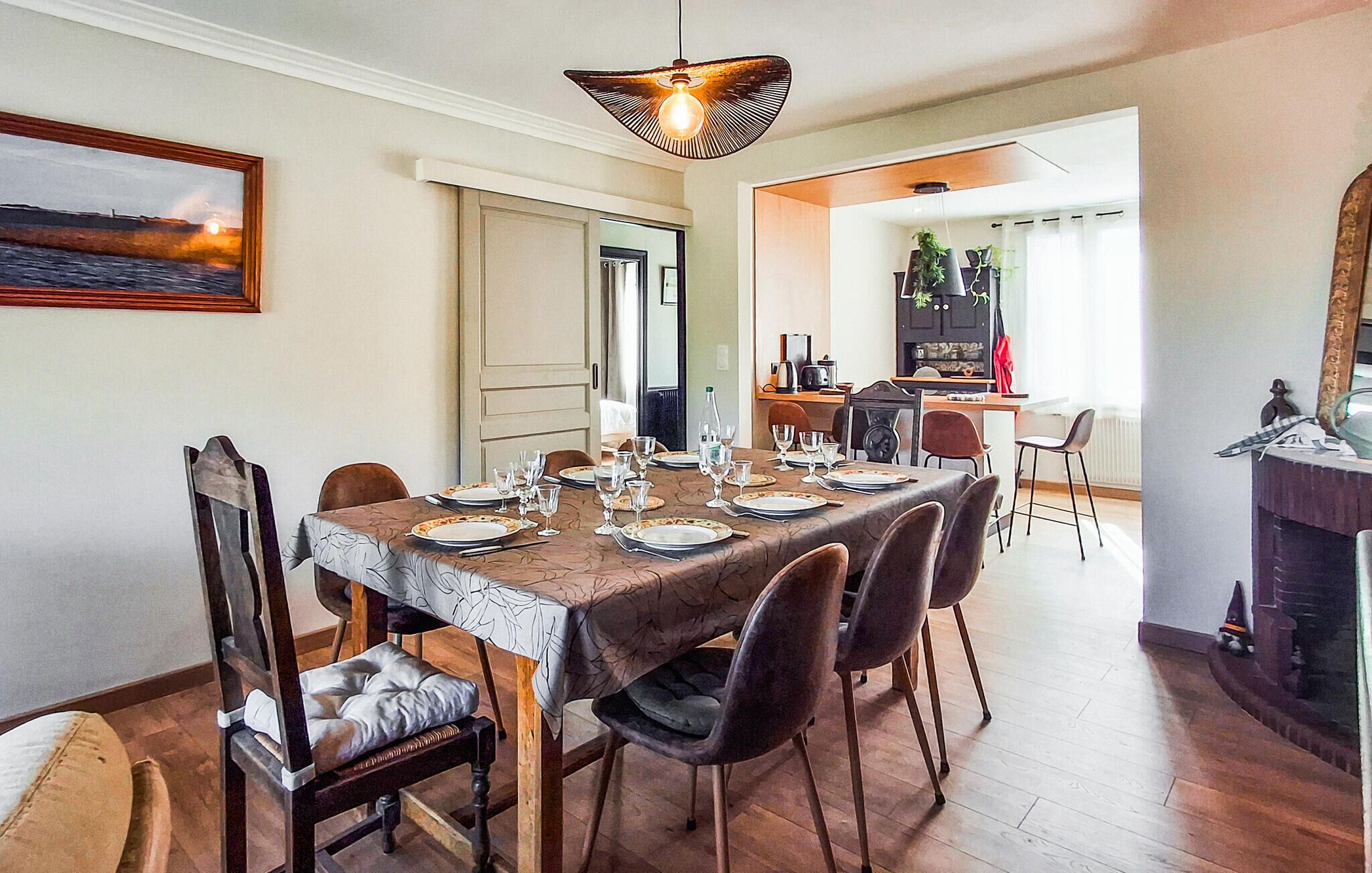 A dining area with a large table set for six, chairs, a hanging light fixture, and a painting on the wall.