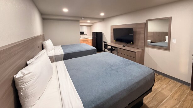 A hotel room with two beds, a desk, a TV, and a small kitchenette in the background.