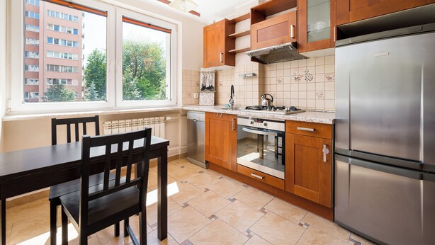 Een keuken met houten kastjes, een roestvrijstalen koelkast, een eettafel met stoelen en een raam met uitzicht op gebouwen en bomen.