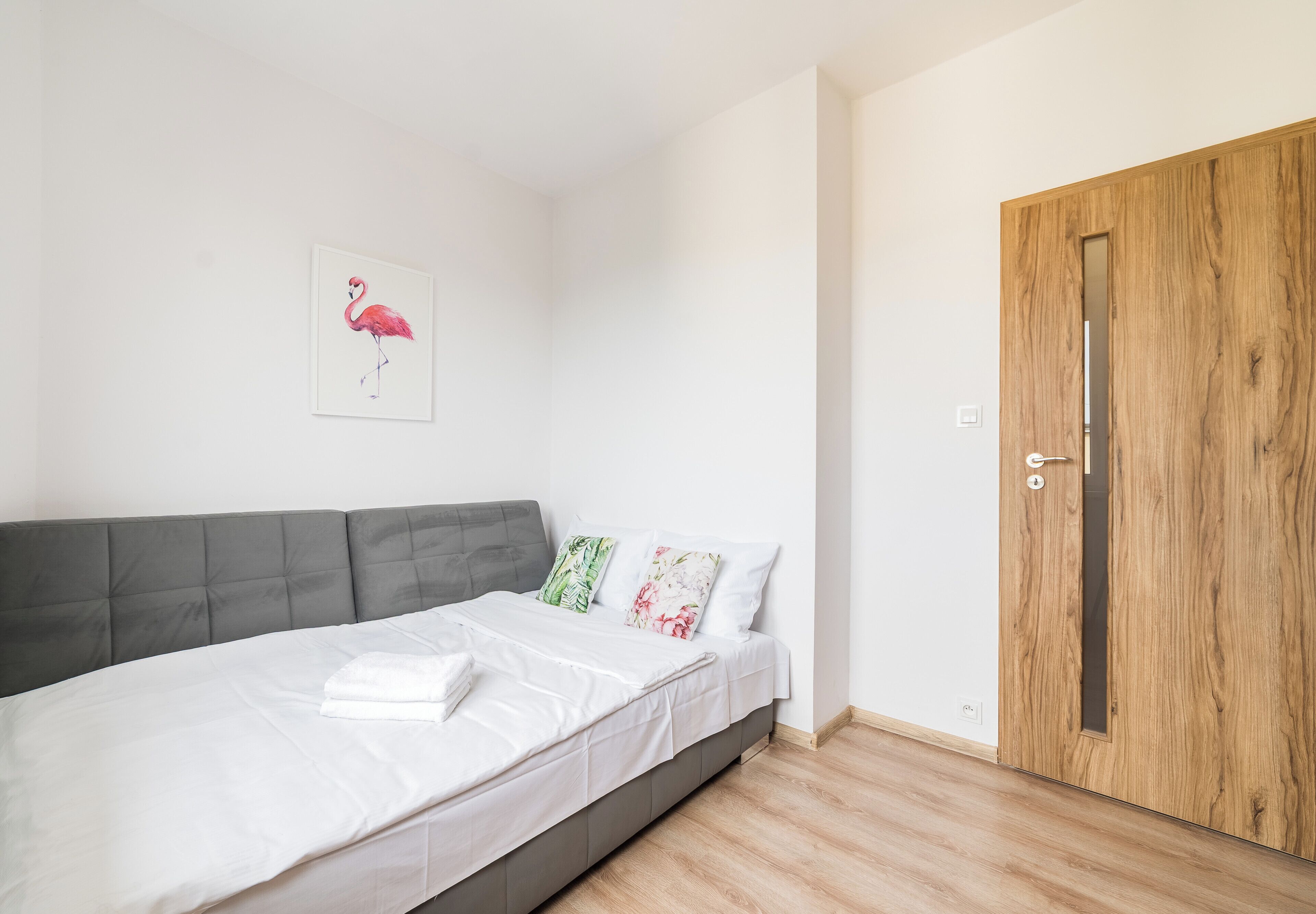 A bedroom with a bed, grey headboard, wooden door, and a wall-mounted artwork of a flamingo.