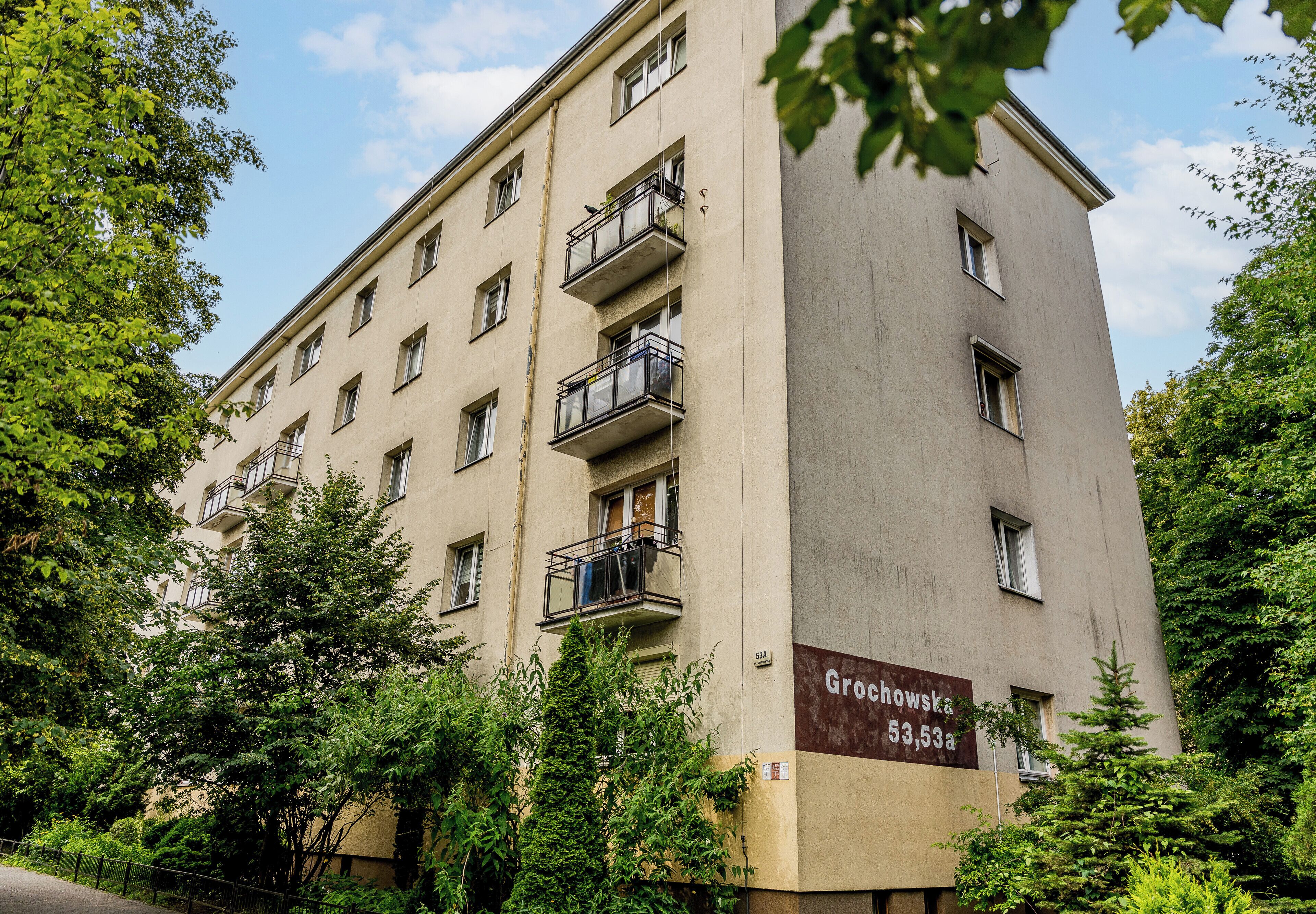 A multi-story building with balconies, a sign indicating 'Grochowska 53,53a,' and surrounding greenery.