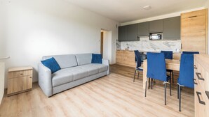 Un salon moderne avec un canapé gris, une table d’appoint en bois et un coin repas comprenant des chaises bleues et une table en bois.