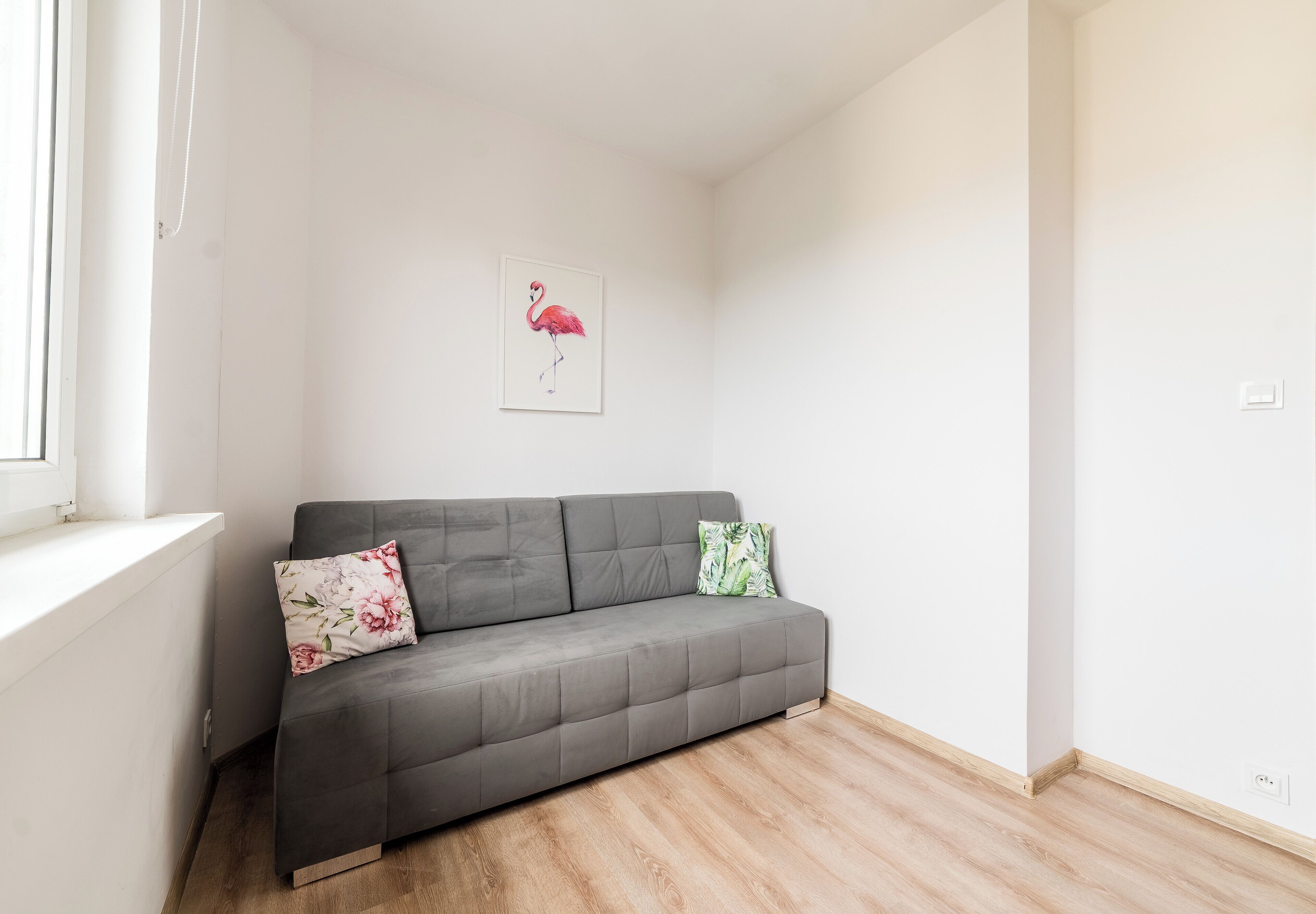Ein graues Sofa mit floralen Kissen, Holzboden, weiße Wand und ein Fenster.