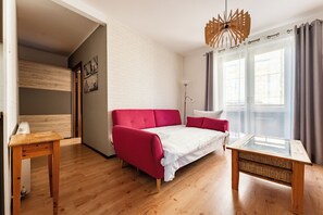 Una habitación con una cama, una mesa de madera, una mesa con cubierta de vidrio, un sofá rojo, una pared blanca y una ventana con cortinas.