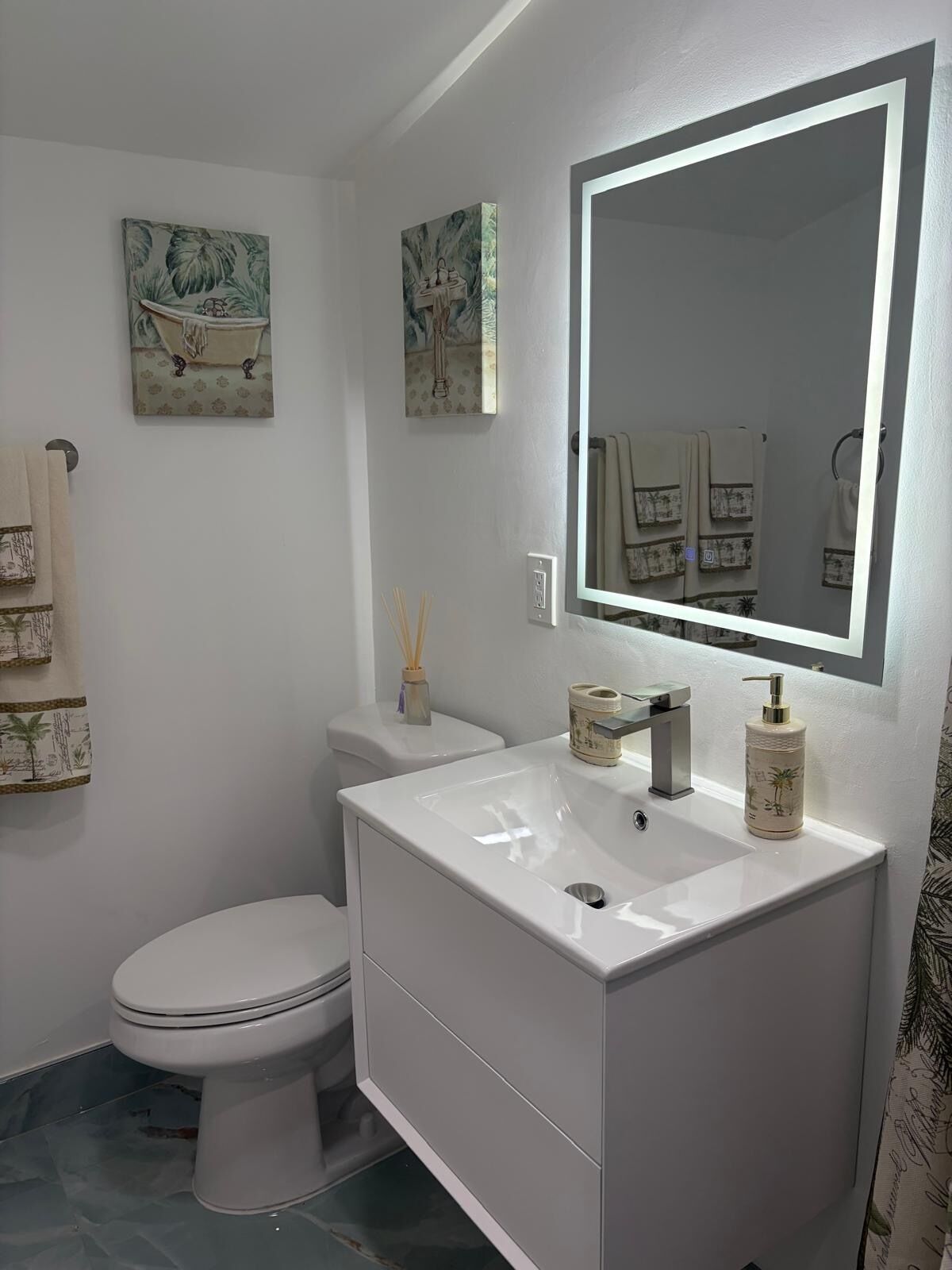 Un baño con lavamanos, inodoro y un espejo grande. Hay dos cuadros en la pared y un perchero con toallas.