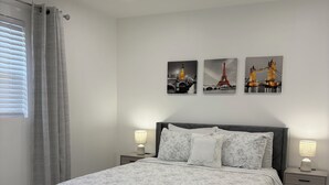 A bedroom with a bed, two bedside tables, a window with curtains, and three wall-mounted pictures.