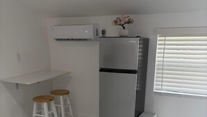 A kitchen with a stainless steel refrigerator, two wooden stools, a white countertop, and a window with blinds.