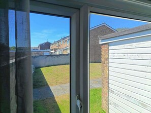Vista a través de una ventana de una zona residencial con un muro de ladrillo, un patio cubierto de césped y una cerca blanca.