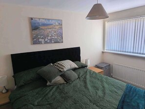 Un dormitorio con cama, un cuadro, una ventana con persianas, un radiador y una mesita de noche con lámpara.