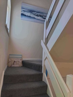 Una escalera con peldaños alfombrados, barandillas blancas y un cuadro de paisaje marino en la pared.