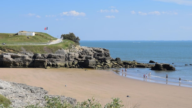 Eine Küstenlandschaft mit Sandstrand, felsigen Klippen und einem grasbewachsenen Hügel mit einer Bebauung.