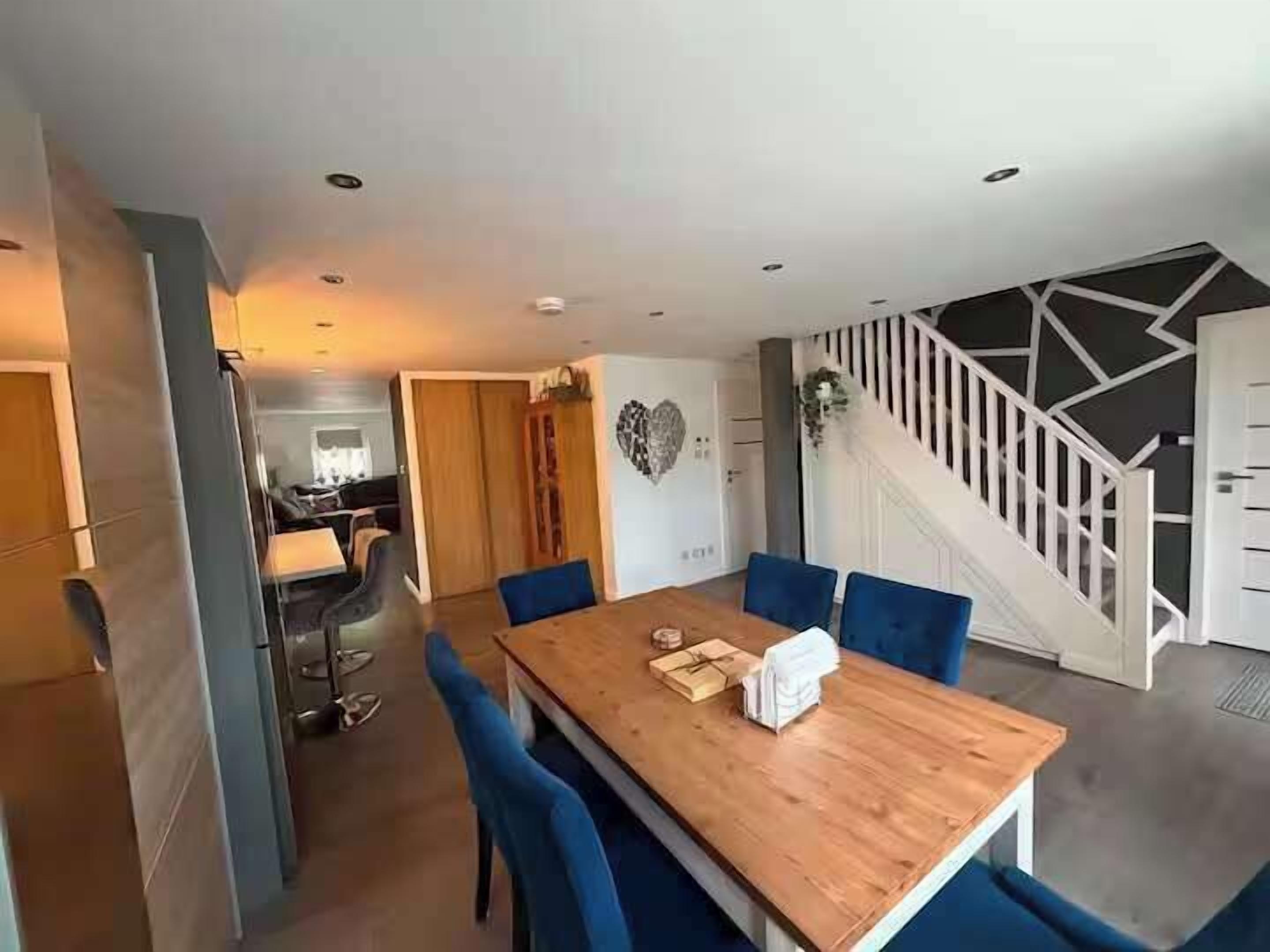 A dining area with a wooden table and blue chairs, a staircase with a geometric design, and a kitchen in the background.