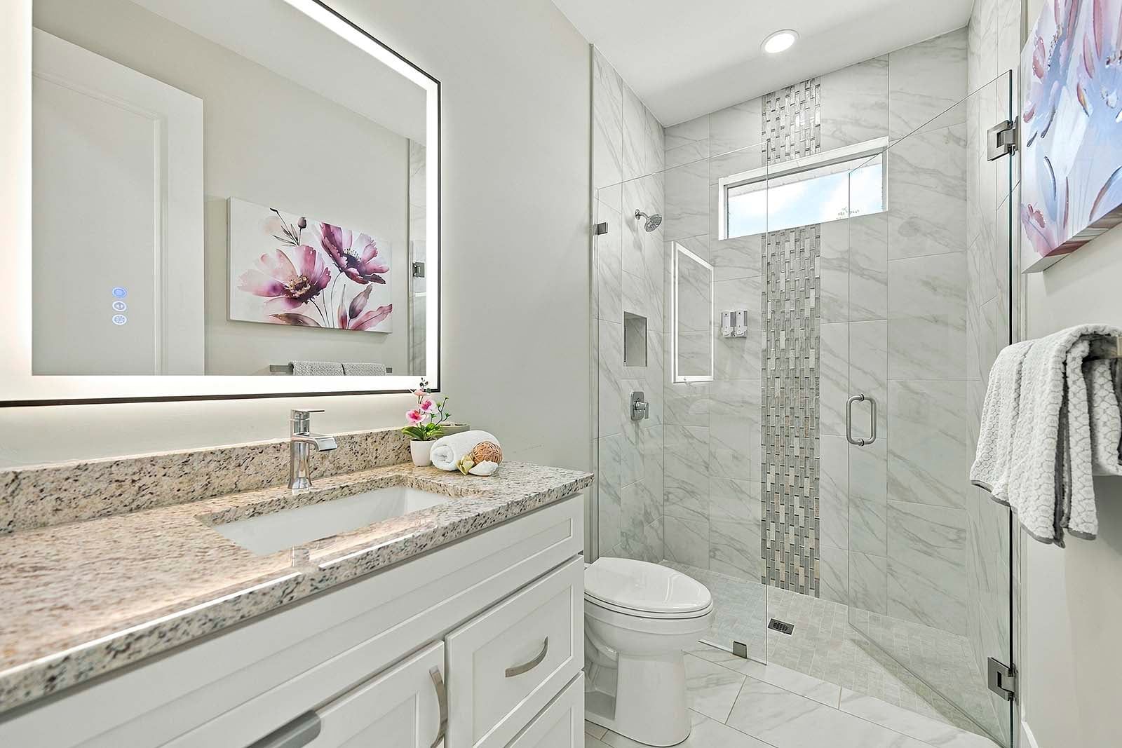 A modern bathroom with a granite countertop, a white toilet, a glass-enclosed shower, and a large mirror with a floral artwork.