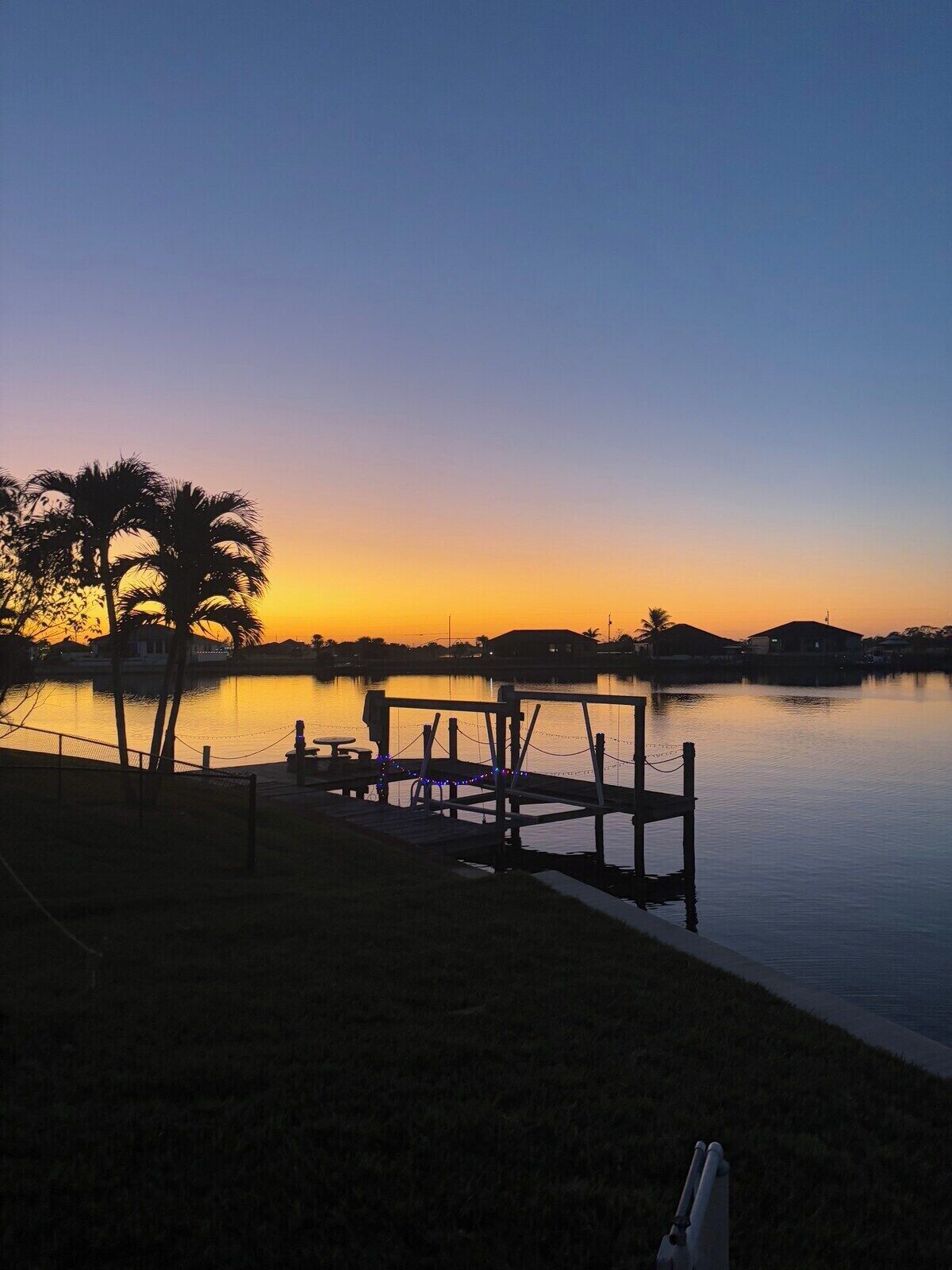 A calm lake at sunset with a dock, palm trees, and a clear sky.