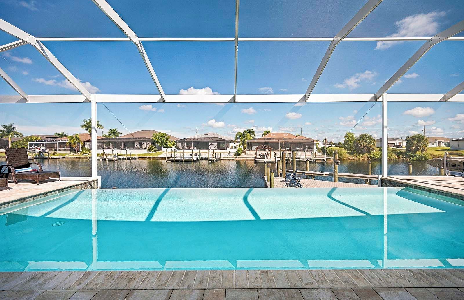 A swimming pool with a wooden deck, a covered area, and a view of houses across the water.