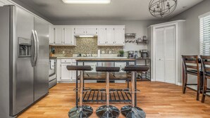 A modern kitchen with white cabinets, a stainless steel refrigerator, a wooden bar stool area, and a chandelier. - Cozy Urban Retreat (Pensacola)