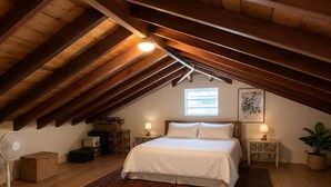 A bedroom with a sloped ceiling, a bed with white bedding, two bedside tables with lamps, a standing fan, a window, and a potted plant.