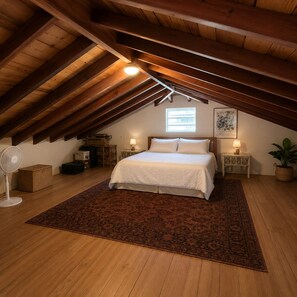 Un dormitorio con techo inclinado, una cama con ropa de cama blanca, dos mesitas de noche con lámparas, un ventilador de pie, una ventana y una planta en maceta.