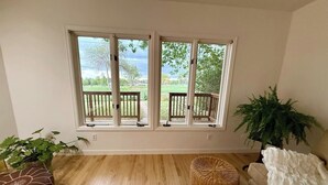 A room with a wooden floor, a wicker ottoman, a white sofa, a potted plant, and a window with a view of a garden. - Sunny Retreat with Stunning Golf Course Views (Centennial)