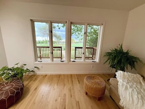 A room with a wooden floor, a wicker ottoman, a white sofa, a potted plant, and a window with a view of a garden.